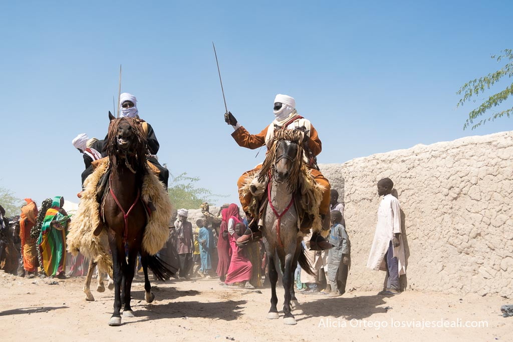 two kanembou horsemen with turbans and swords held aloft followed by a group of people