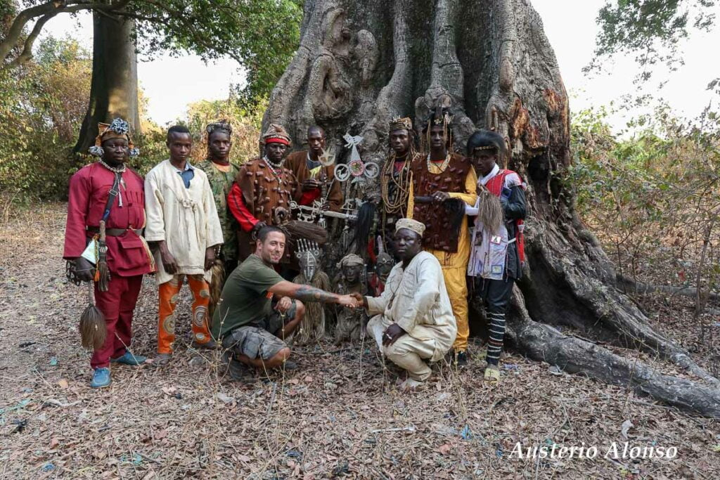 Alonso posing with dozo hunters and their fetishes with a big tree behind him.